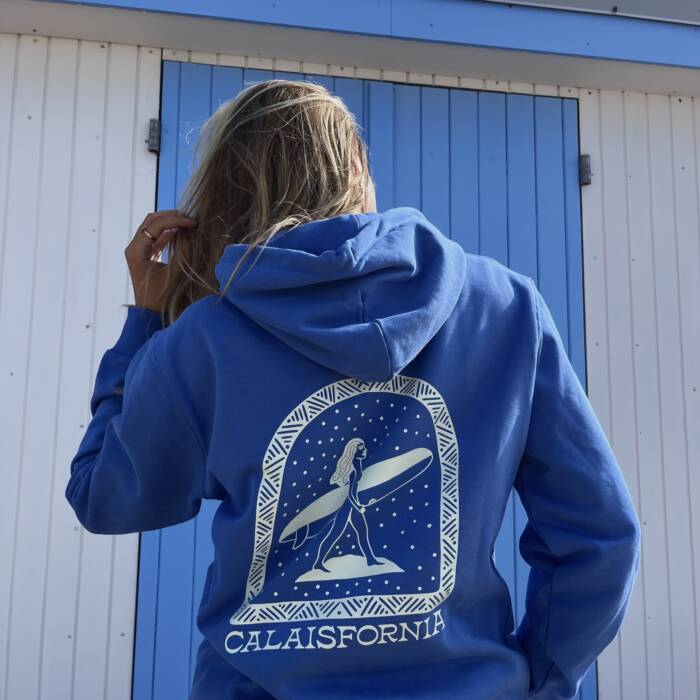 Sweatshirt bleu de la marque calaisienne “Calaisfornia” avec une illustration rétro d’une surfeuse, porté devant les cabines de plage iconiques de Calais. Mélange d’inspiration californienne et maritime, mettant en valeur l’esprit côtier et local de la région.