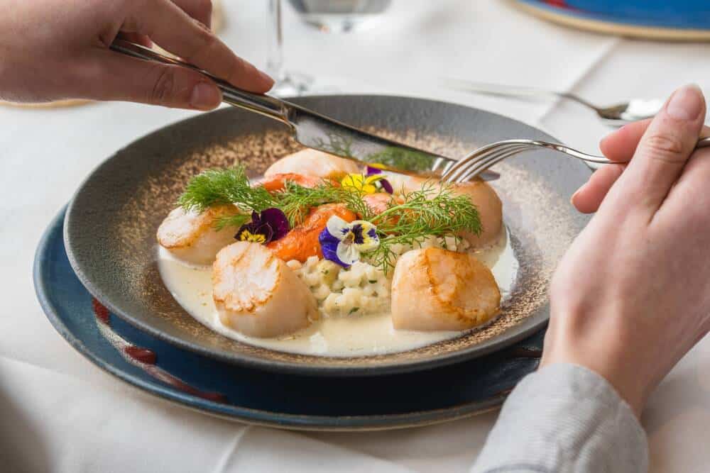 Assiette gastronomique de Noix de Saint-Jacques poêlées servie au restaurant L’Aquar’aile à Calais. Accompagnée d’un risotto crémeux, d’aneth frais et de fleurs comestibles pour une présentation élégante et raffinée. Cuisine locale mettant en avant les produits de la mer.