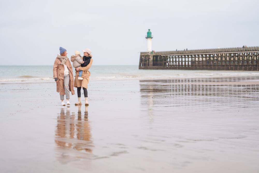 Une famille en promenade hivernale sur la plage de Calais, près de la jetée et de son phare emblématique. Le sable humide reflète le ciel gris, créant une scène paisible et naturelle, parfaite pour un moment de détente en bord de mer en toute simplicité.