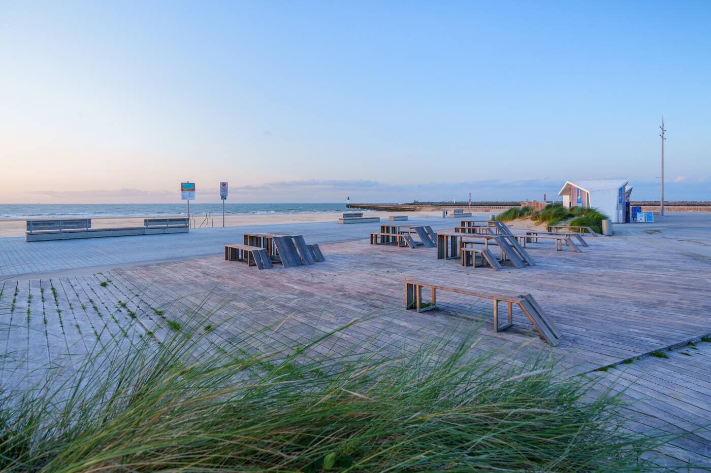 Aménagements familiaux sur la plage de Calais, comprenant des bancs et tables en bois installés sur une terrasse en bord de mer. L’espace, entouré de dunes et offrant une vue dégagée sur l’horizon, invite à la détente et aux moments de convivialité en plein air.