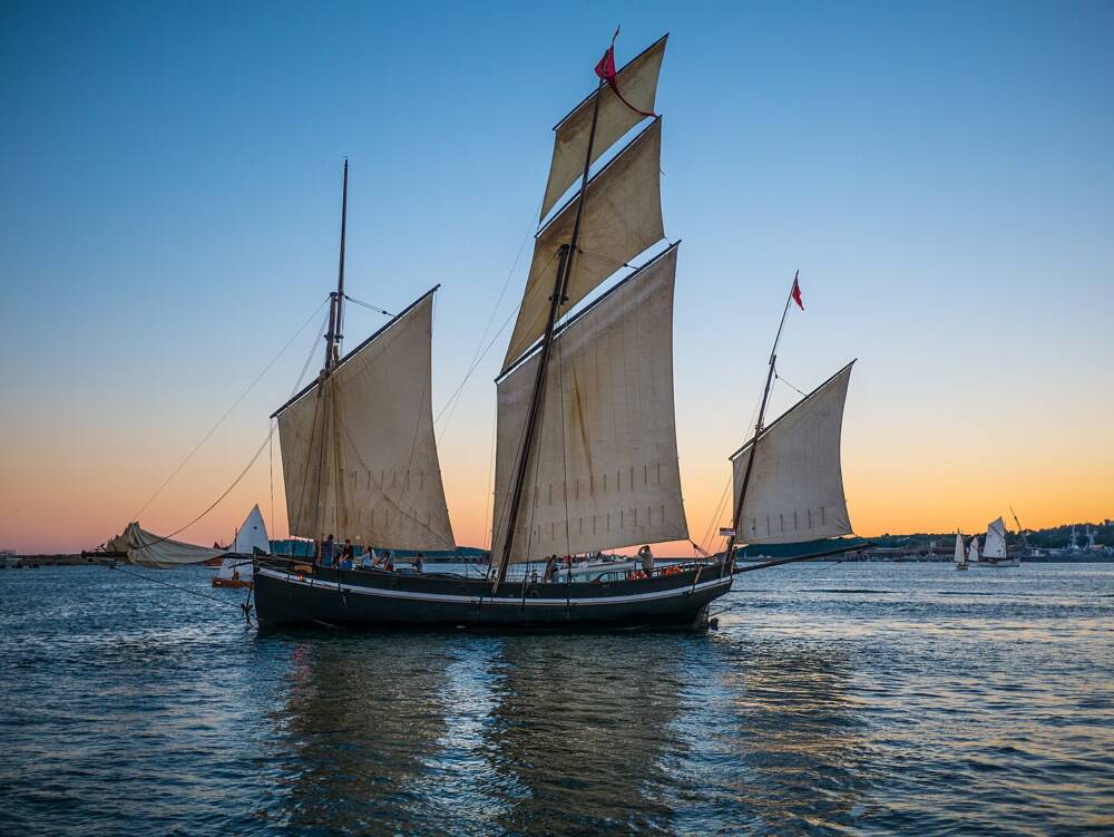 Le Grayhound, goélette trois-mâts noire aux voiles beiges, naviguant sur une mer calme au coucher du soleil.