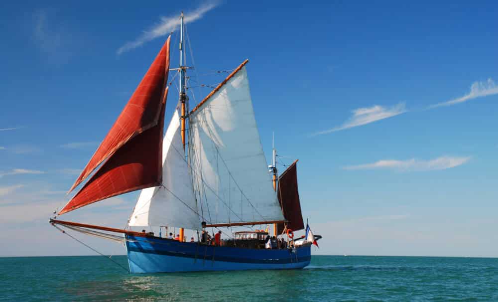 Description alt optimisée SEO : Le Milpat, voilier traditionnel aux voiles rouges et à la coque bleue, naviguant sous un ciel dégagé. Ce navire emblématique sera présent à Escale à Calais, offrant une immersion dans l’histoire maritime et la navigation à l’ancienne.