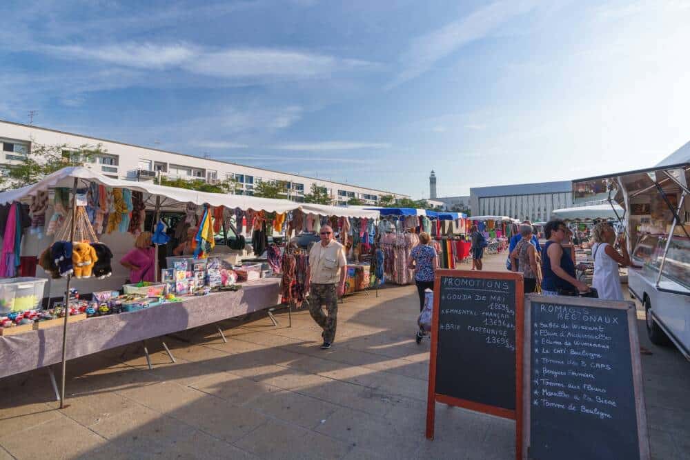 Marché ensoleillé sur la place d’Armes de Calais, proposant vêtements, artisanat et produits locaux. Une ambiance chaleureuse où habitants et touristes profitent des étals variés, avec en arrière-plan le Phare et l'architecture moderne de la Halle couverte.